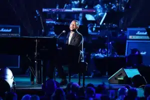 A person is playing a grand piano on stage, illuminated by stage lights, with a band visible in the background.