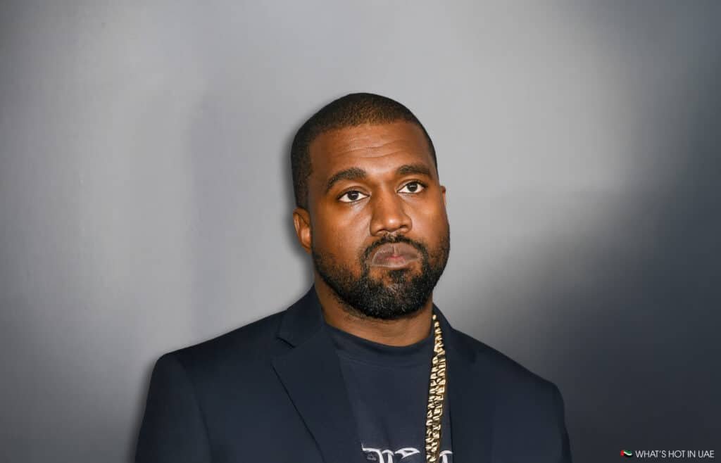 Kanye West with a serious expression, wearing a dark blazer and a gold chain, standing against a plain background.