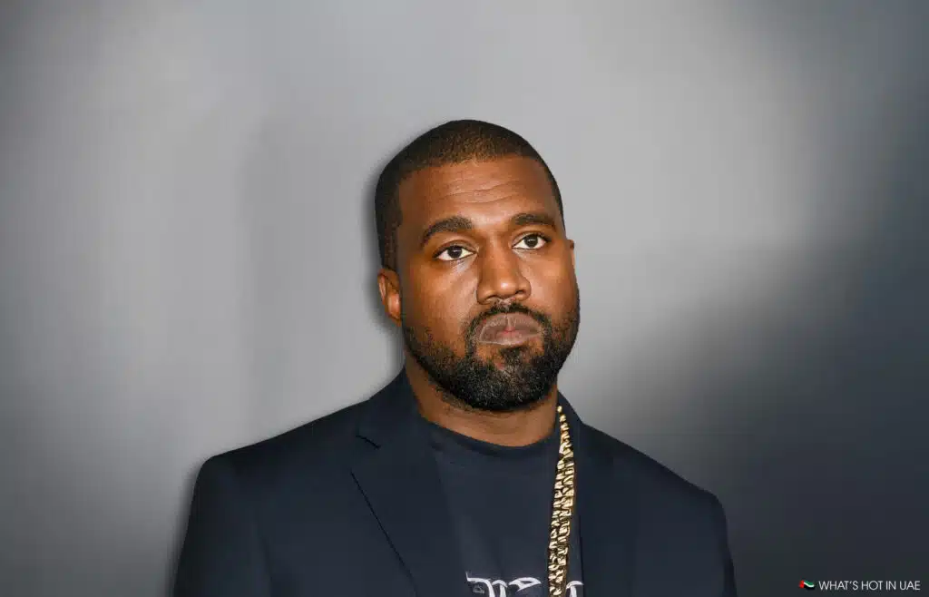 Kanye West with a serious expression, wearing a dark blazer and a gold chain, standing against a plain background.
