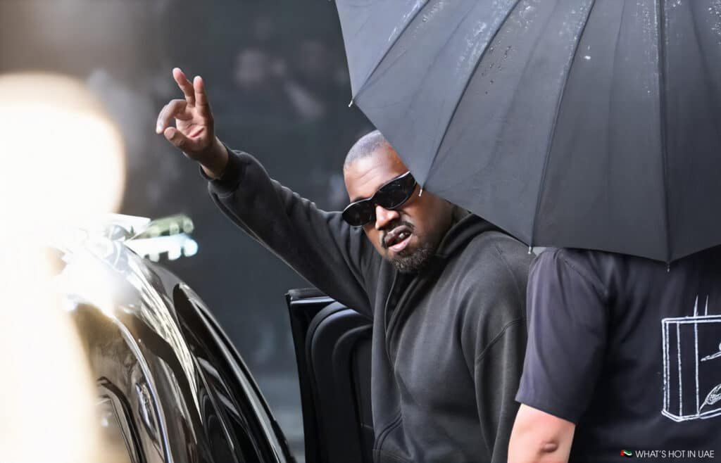 Kanye West wearing sunglasses and a dark hoodie makes a hand gesture while stepping out of a vehicle, partially covered by a large umbrella.