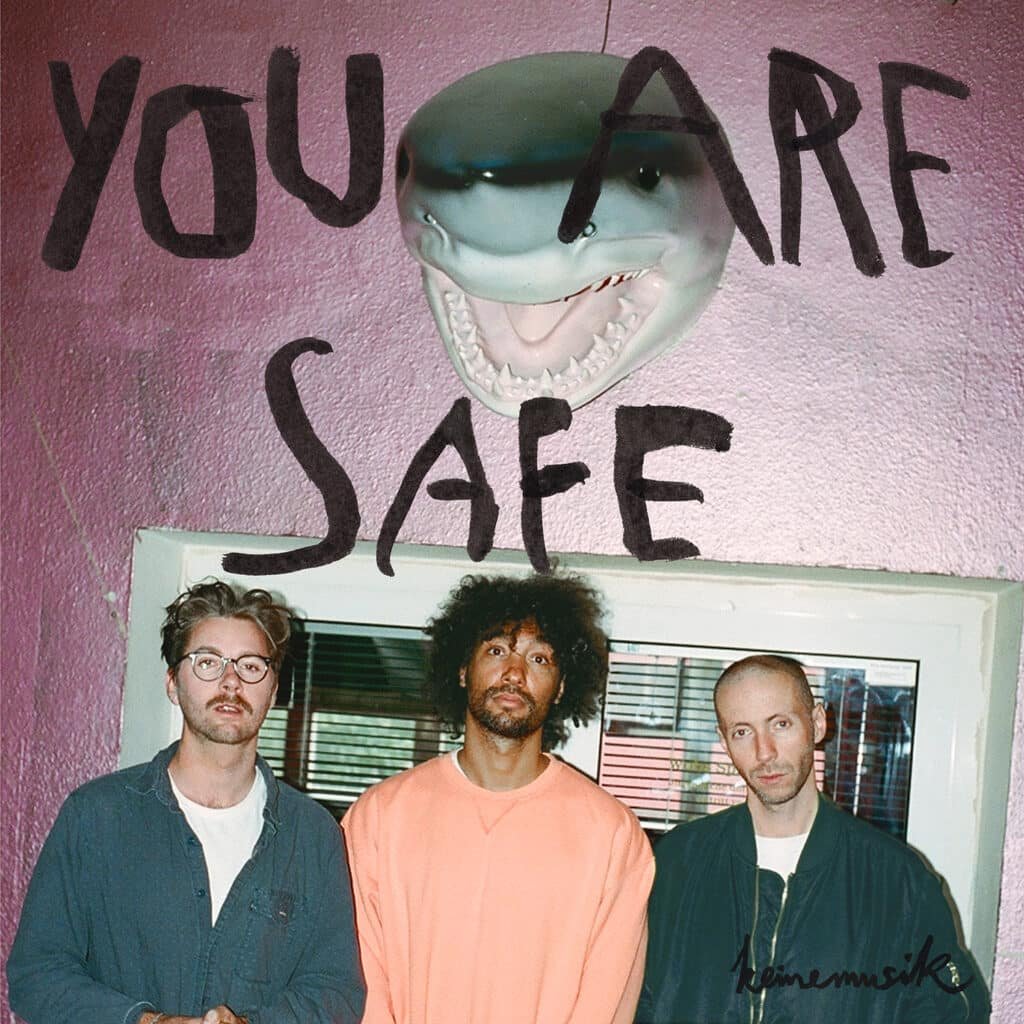 Three men stand in front of a pink wall with a mounted shark head above them and bold text reading "YOU ARE SAFE."