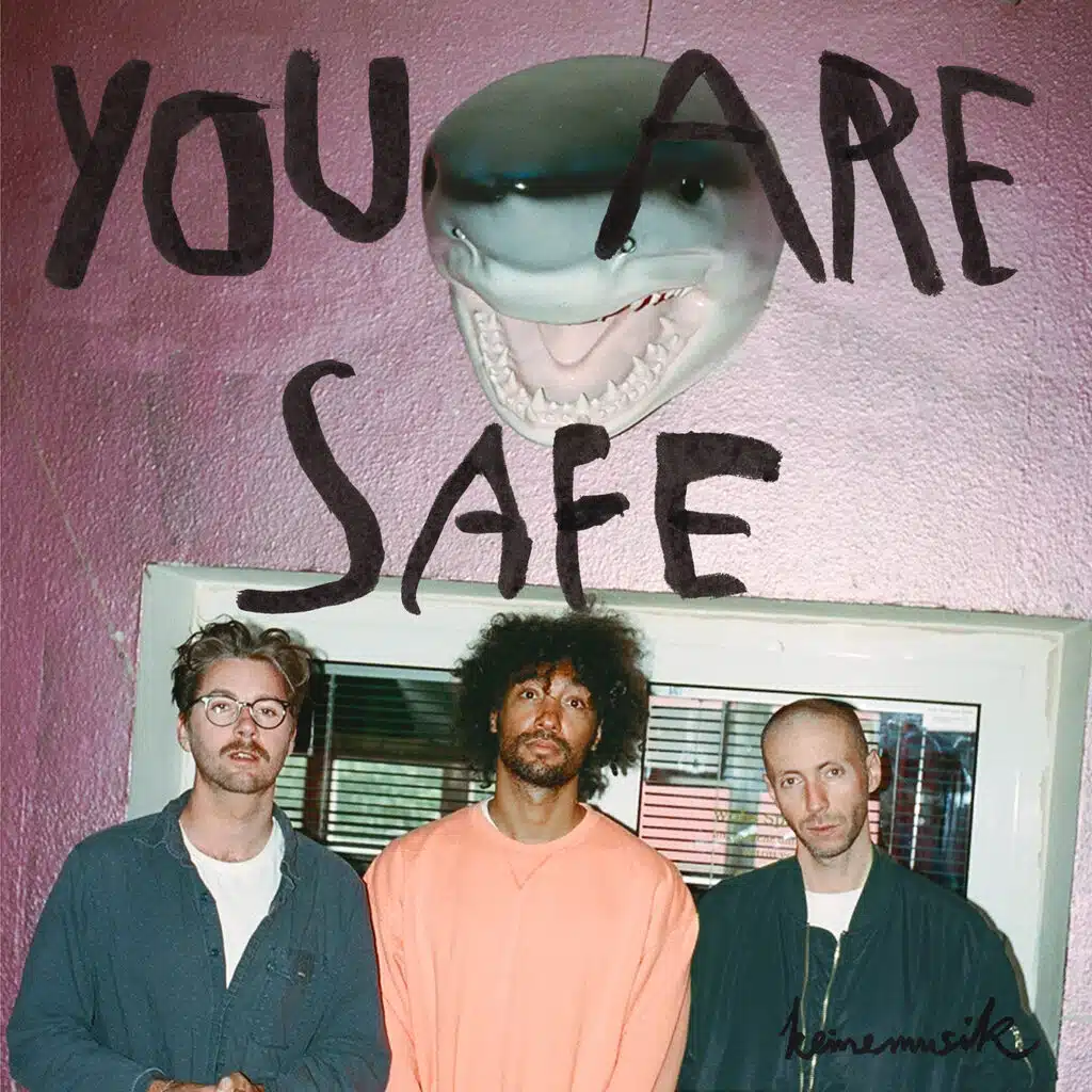Keinemusik stand in front of a pink wall with a mounted shark head above them and bold text reading "YOU ARE SAFE."