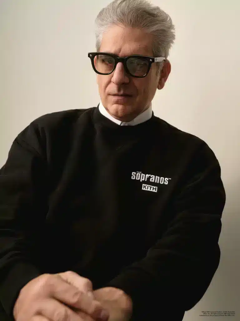 A man wearing glasses and a black sweatshirt with "Kith x The Sopranos" printed on it, sitting against a neutral background.