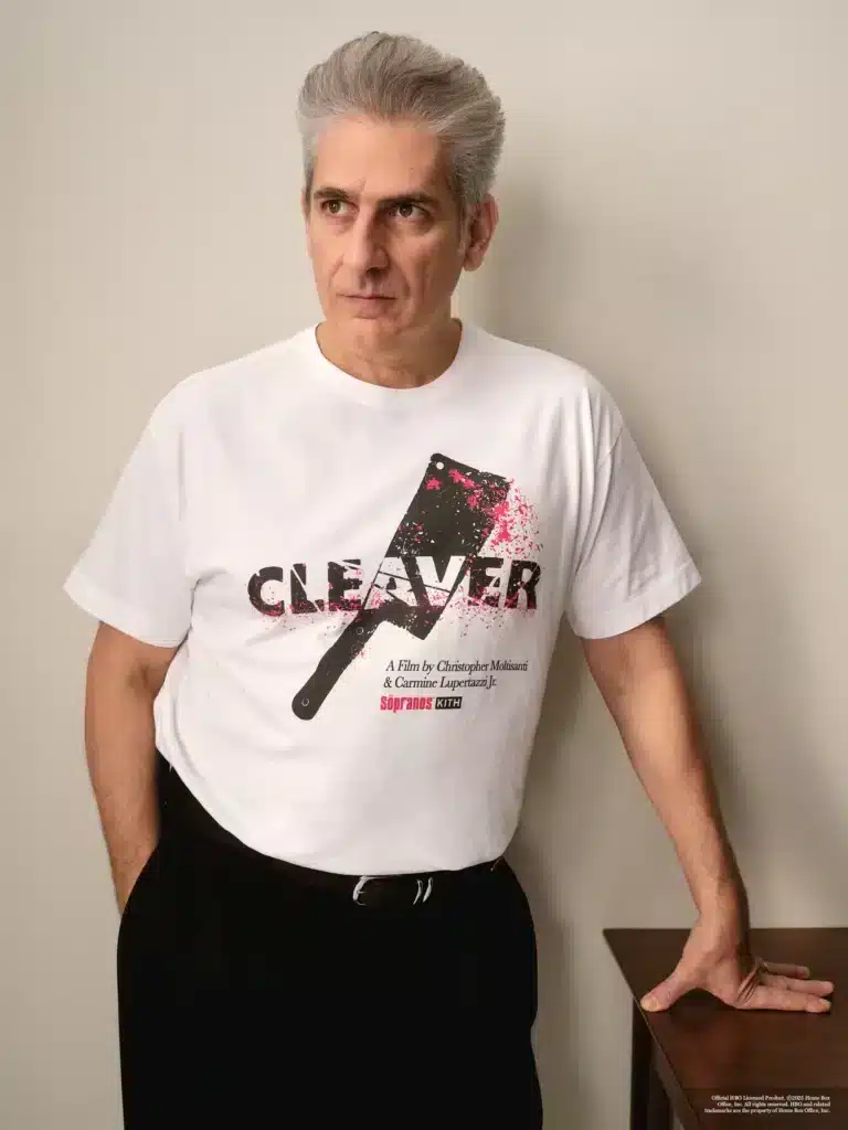 Person wearing a white t-shirt with a graphic design featuring a cleaver and text, leaning against a table.