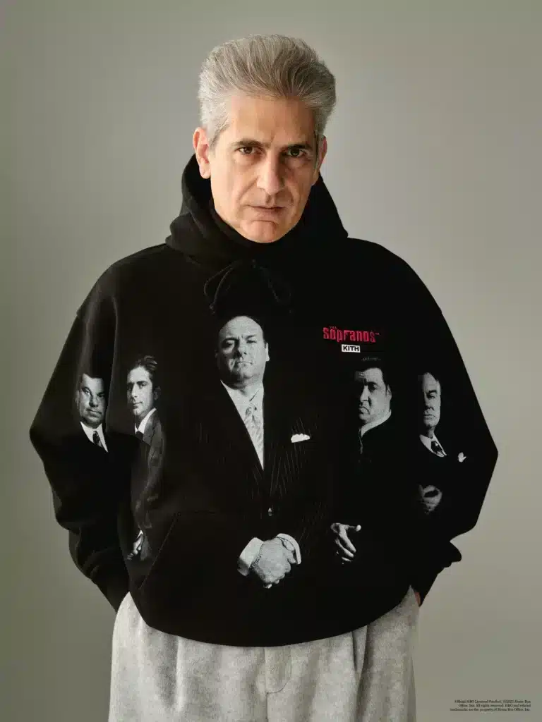 A man with gray hair wearing a black Kith x The Sopranos hoodie featuring images and text related to a TV show, standing against a neutral background.