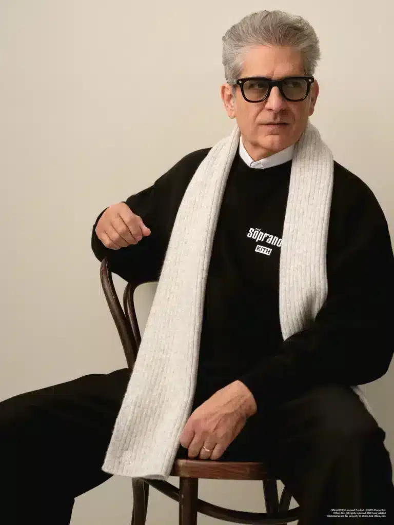 A man with glasses and gray hair sits on a chair wearing a black sweater with "Kith x The Sopranos" text and a long white scarf.