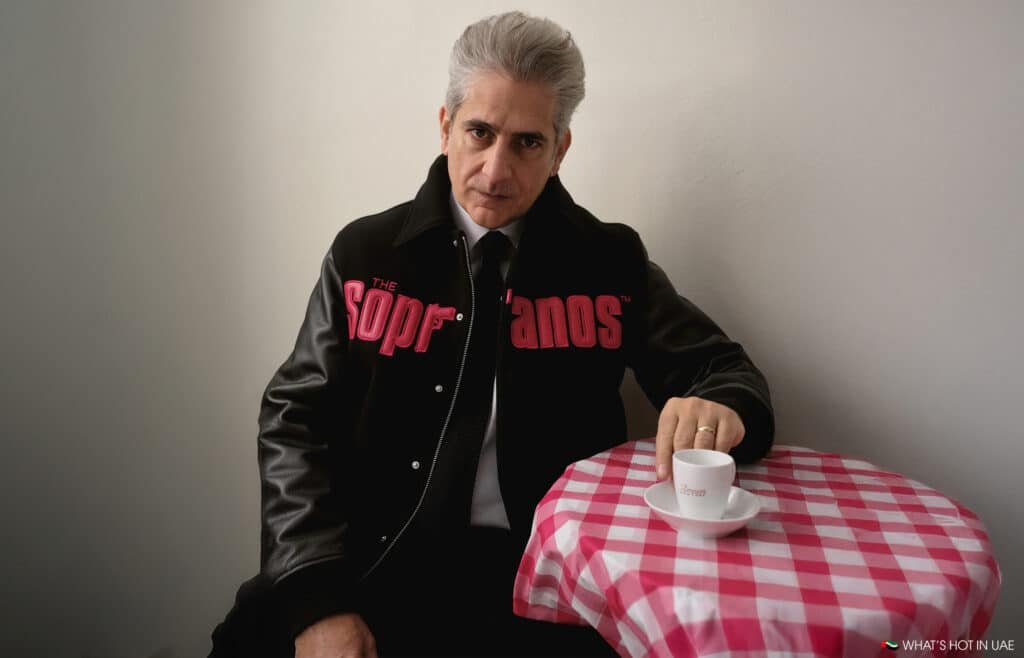 A man with gray hair wearing a black jacket with "The Sopranos" logo sits at a red-checkered table with an espresso cup.