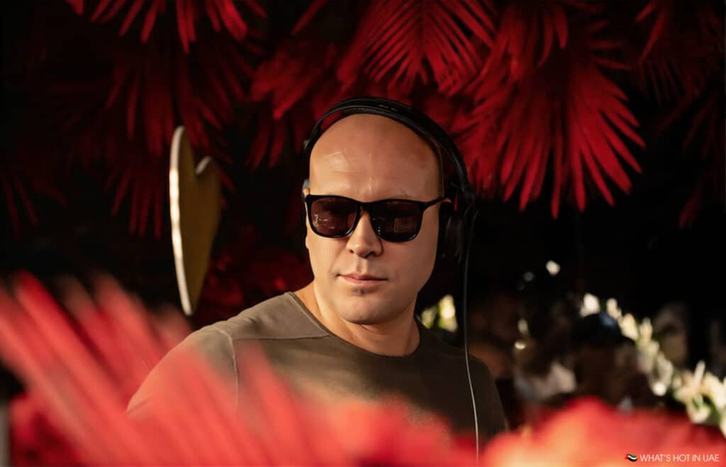 Marco Carola wearing sunglasses and headphones, standing in front of red foliage, possibly at a music event.