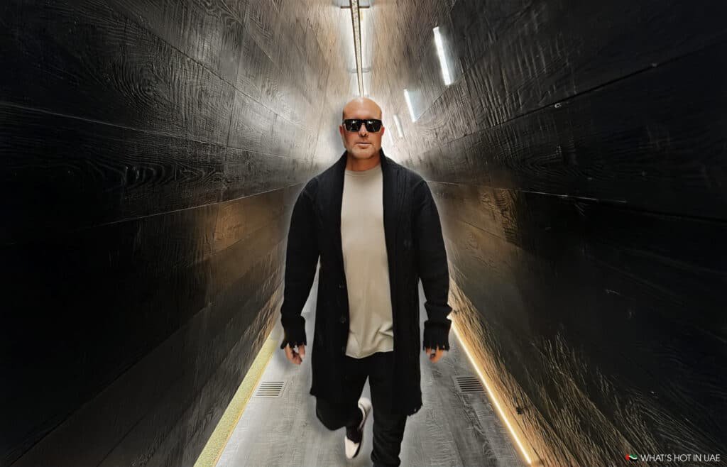 A person wearing sunglasses and a black coat walking through a dark, narrow hallway with illuminated walls.