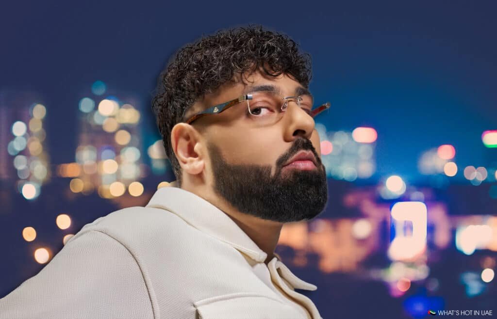 A man with MAYBACH Eyewear glasses and a beard, wearing a light-colored jacket, looking over his shoulder with a blurred cityscape in the background.