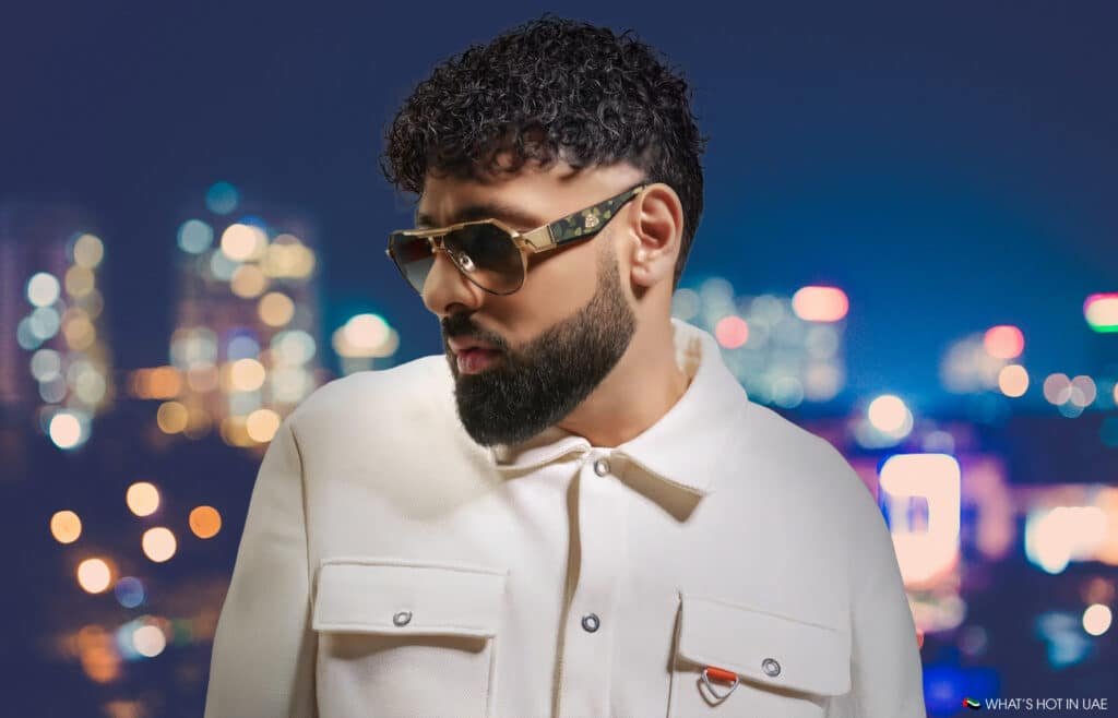 Person with curly hair and a beard wearing sunglasses and a white jacket, standing against a blurred cityscape background at night.