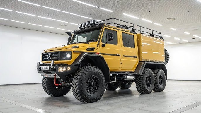 A yellow Mercedes-Benz G-Class 6x6 vehicle with large off-road tires and a rugged, customized body in a showroom setting.