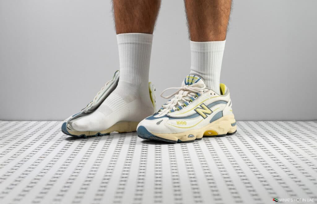 A person wearing mismatched sneakers, one a New Balance shoe, on a patterned floor.