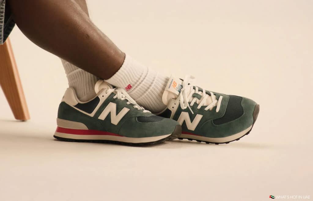 A person wearing green and white New Balance sneakers with white socks, sitting on a wooden chair.