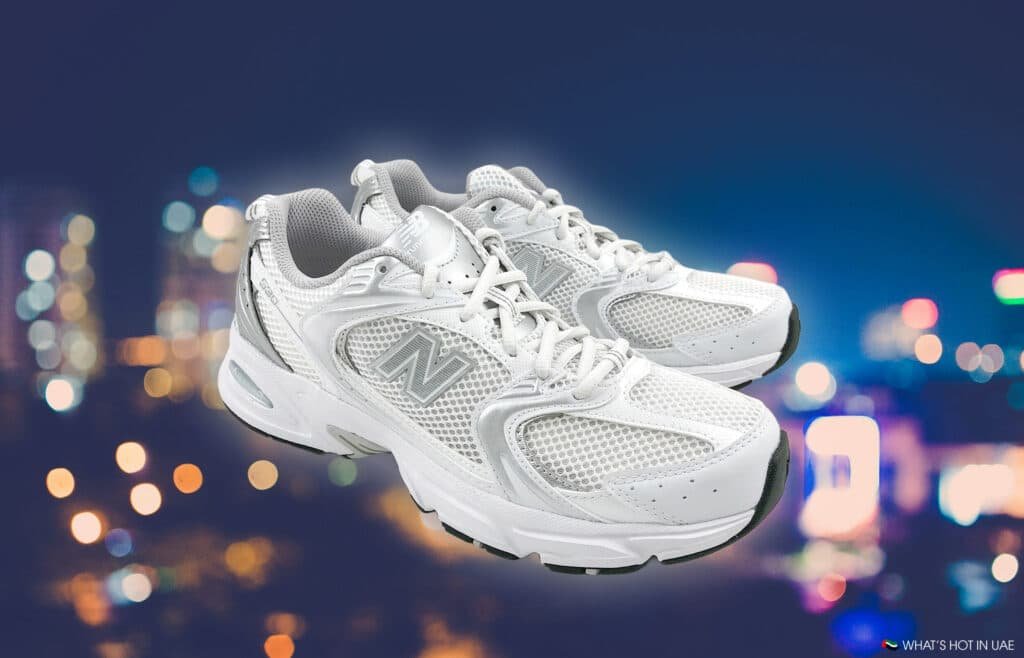 A pair of white athletic shoes with mesh panels and gray detailing, displayed against a blurred city lights background.