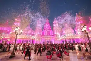 A vibrant festival scene with performers in colorful costumes and drummers in front of an ornate, pink-lit building.