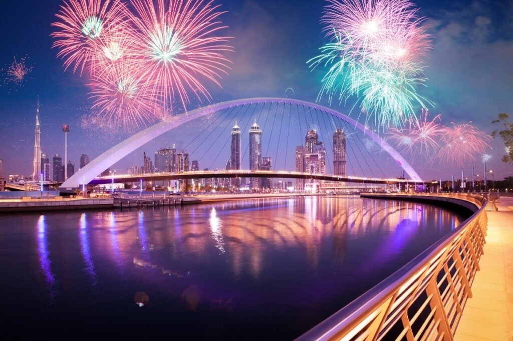 A vibrant NYE 2026 fireworks display over a modern cityscape with a lit-up arch bridge reflecting in the water.