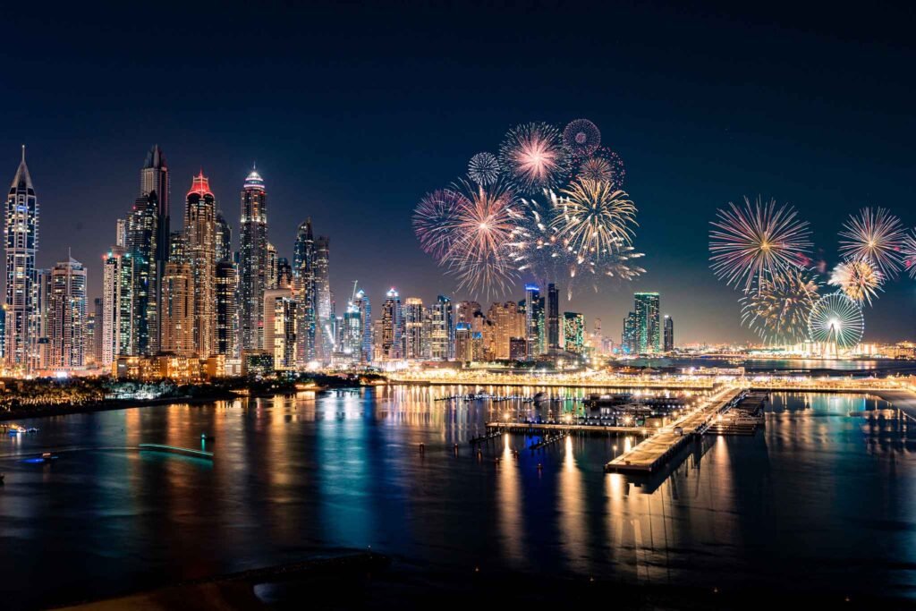 A nighttime cityscape with skyscrapers is illuminated by colorful fireworks over a waterfront reflecting the vibrant lights.