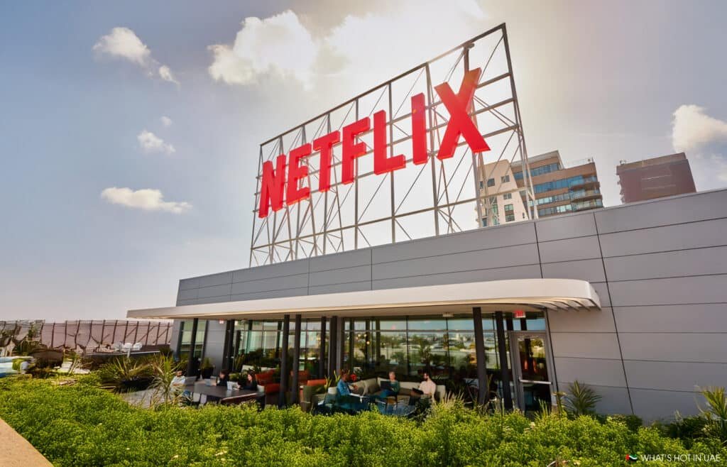 A rooftop scene featuring a large red "Netflix" sign atop a modern building, with people sitting and talking on a terrace below surrounded by green plants.