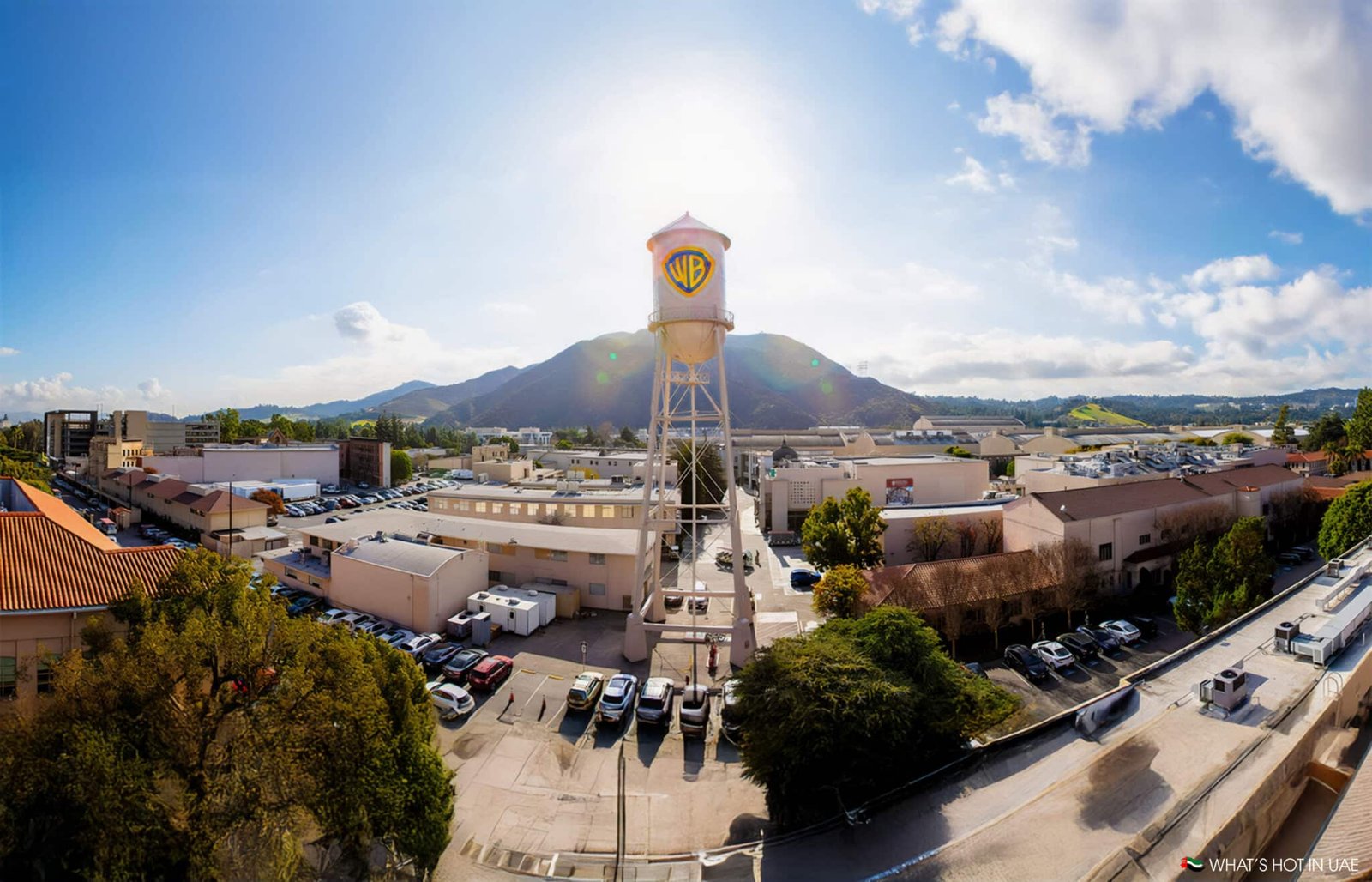 A view of the Warner Bros. studio lot featuring a prominent water tower with the WB logo, surrounded by various buildings and parking areas, set against a backdrop of rolling hills under a partly cloudy sky.