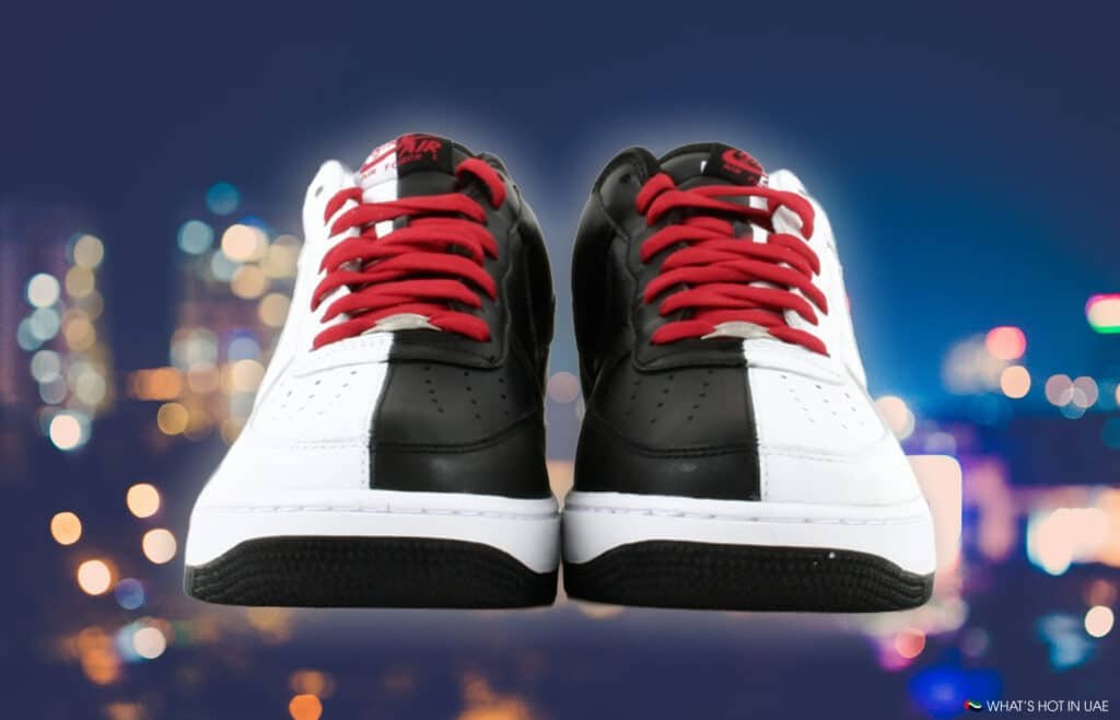 A pair of black and white Nike Air Force 1 Low 'Scarface' sneakers with red laces against a blurred cityscape background.