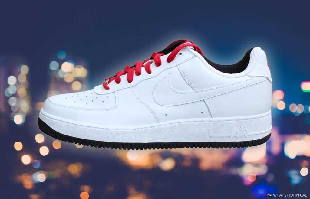 A white sneaker with red laces and a black sole, featuring the Nike swoosh logo, set against a blurred cityscape background.