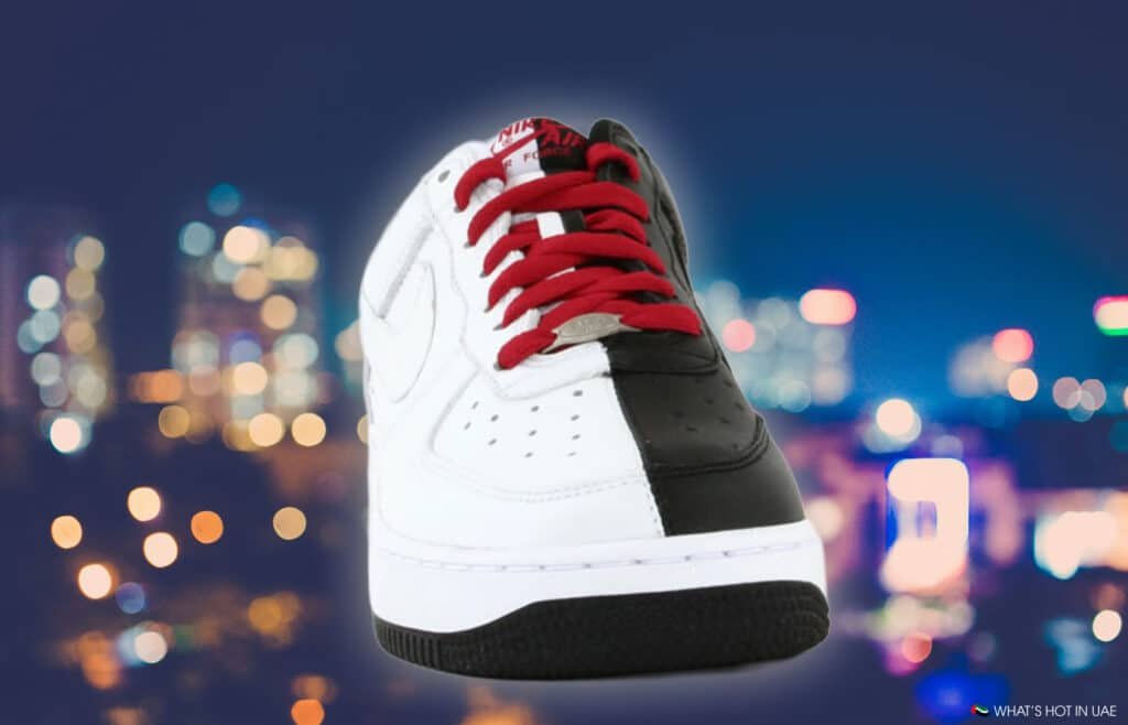 A sneaker with a split design of white and black leather, featuring red laces, against a blurred city lights background.