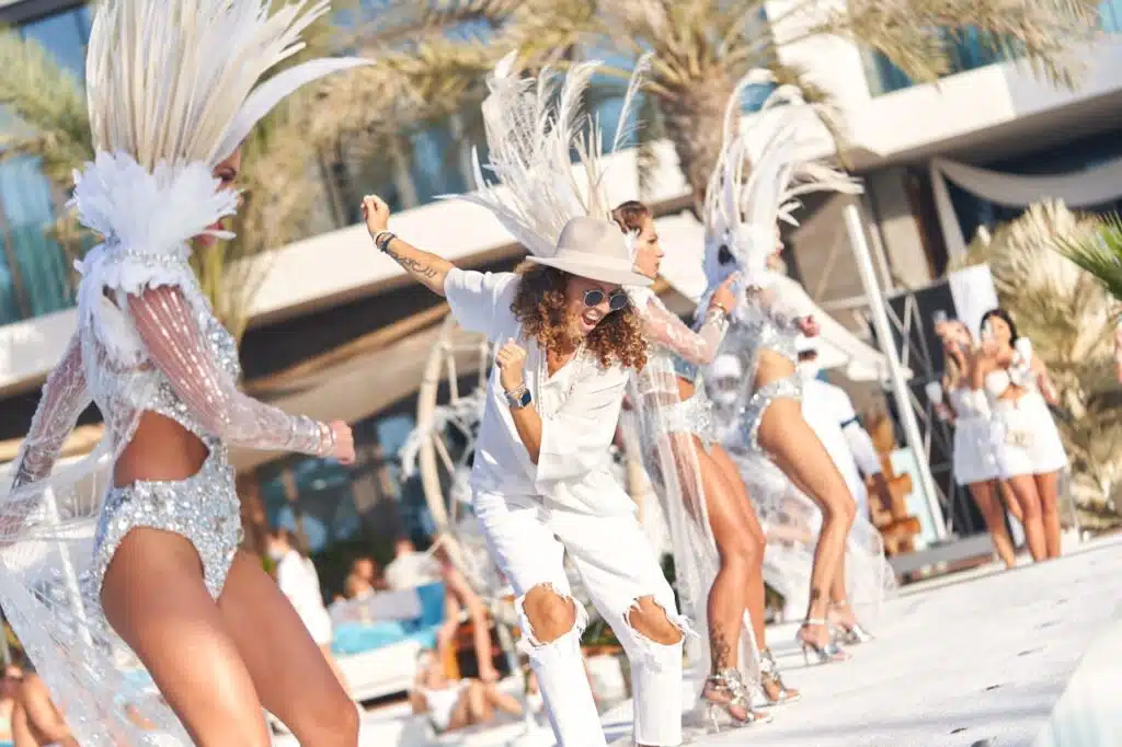 A group of performers dressed in white and silver costumes with feathered headpieces dance energetically outdoors, with palm trees and a modern building in the background.
