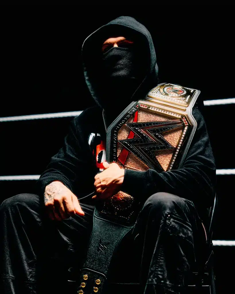 A person wearing a hooded jacket and mask sits in a dimly lit space, holding a championship wrestling belt.