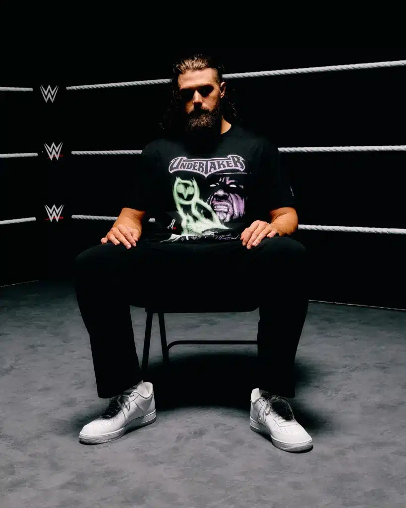 OVO x WWE: A person with long hair and a beard sits on a chair in a wrestling ring, wearing a black Undertaker graphic t-shirt and white sneakers.