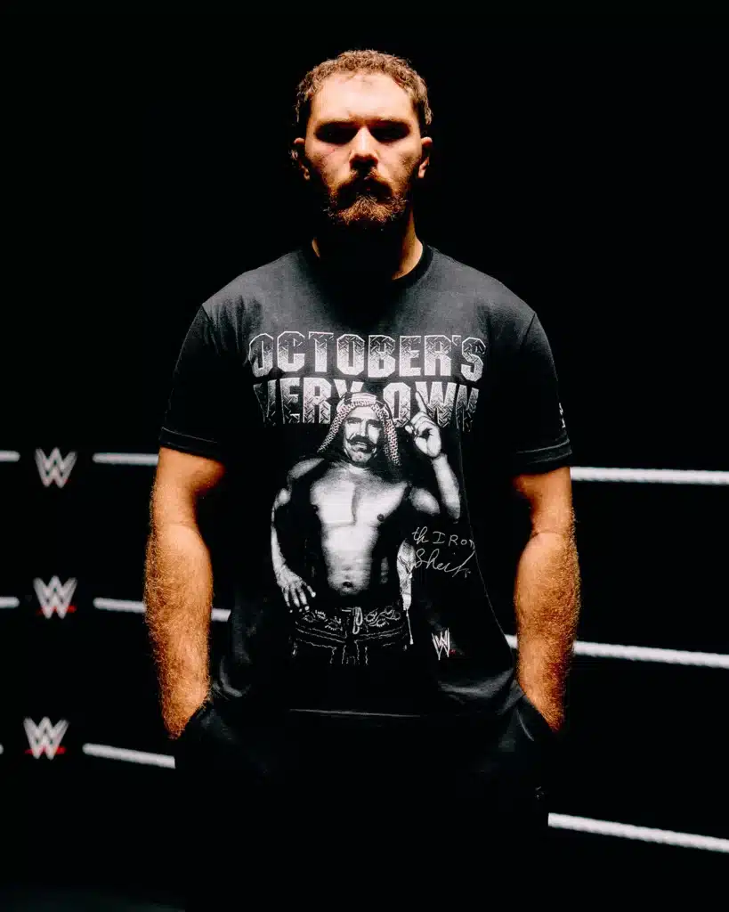 OVO x WWE: Man with a beard wearing a T-shirt featuring a wrestler, standing in a dimly lit wrestling ring with visible ropes and WWE logos.