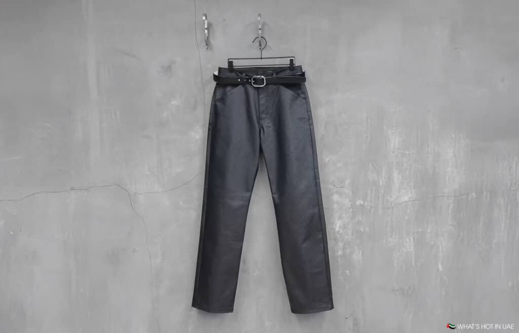 Black leather Omar Afridi x Vuja Dé pants with a belt hanging on a hanger against a textured gray wall.