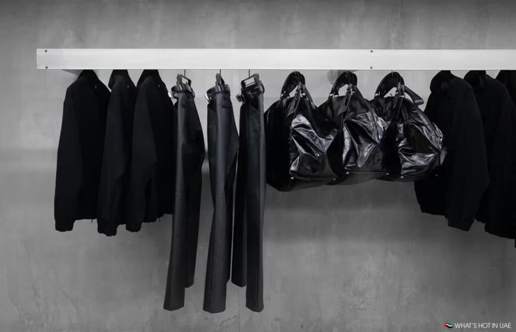 A minimalist display featuring several Omar Afridi x Vuja Dé black jackets, pants, and shiny black bags hanging on a white rack against a concrete wall.