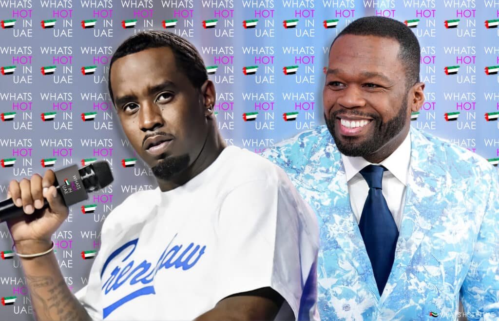 Two men posing together in front of a backdrop with repeated text "What's Hot in UAE" and an image of the UAE flag. One man holds a microphone with the same logo, while the other is smiling and wearing a blue patterned suit.