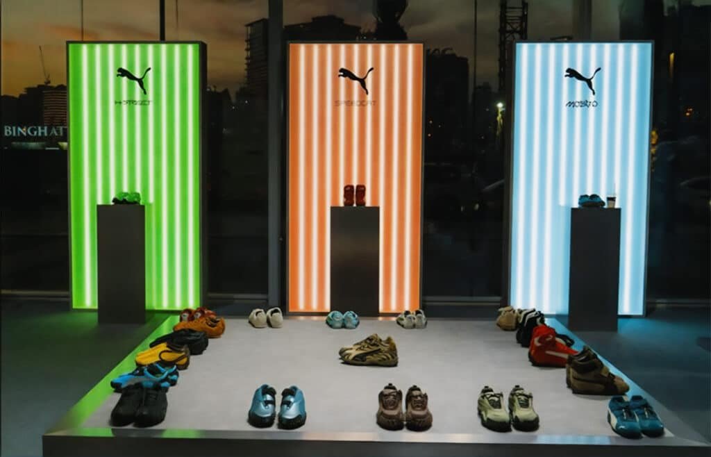 Shoe display featuring various pairs under illuminated panels with Puma and Adidas logos, in a store setting.