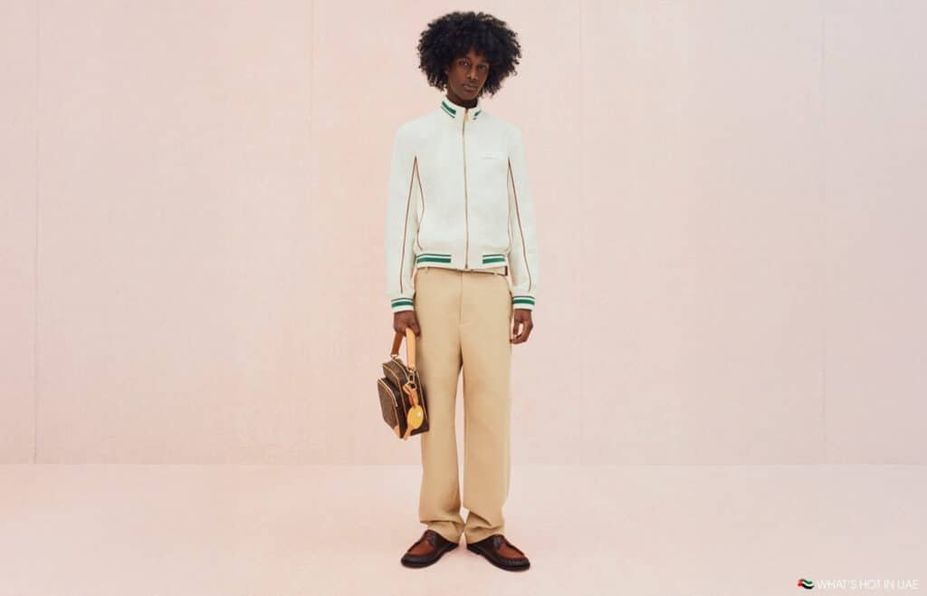 A person standing against a plain backdrop wearing a white jacket with green trim, beige pants, brown shoes, and holding a brown handbag.