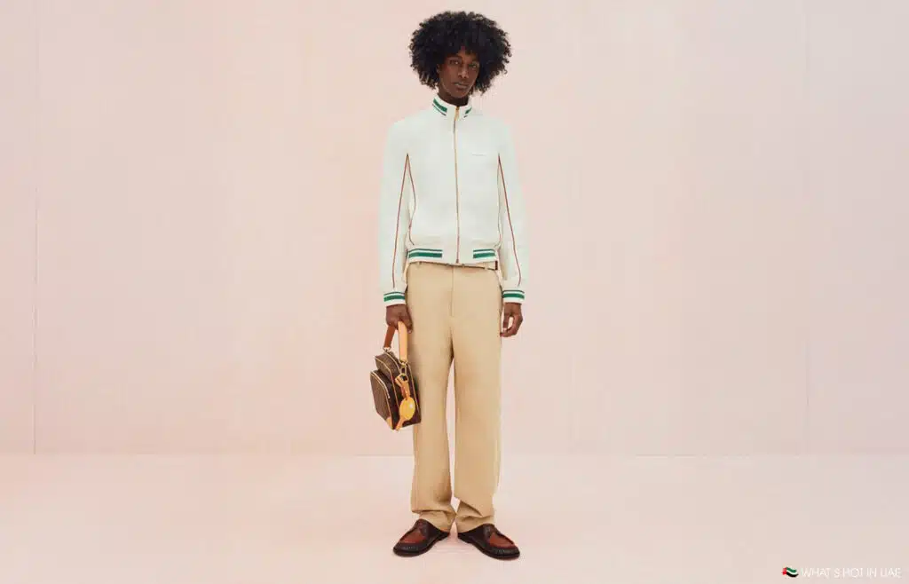 A person standing against a plain backdrop wearing a white jacket with green trim, beige pants, brown shoes, and holding a brown handbag.