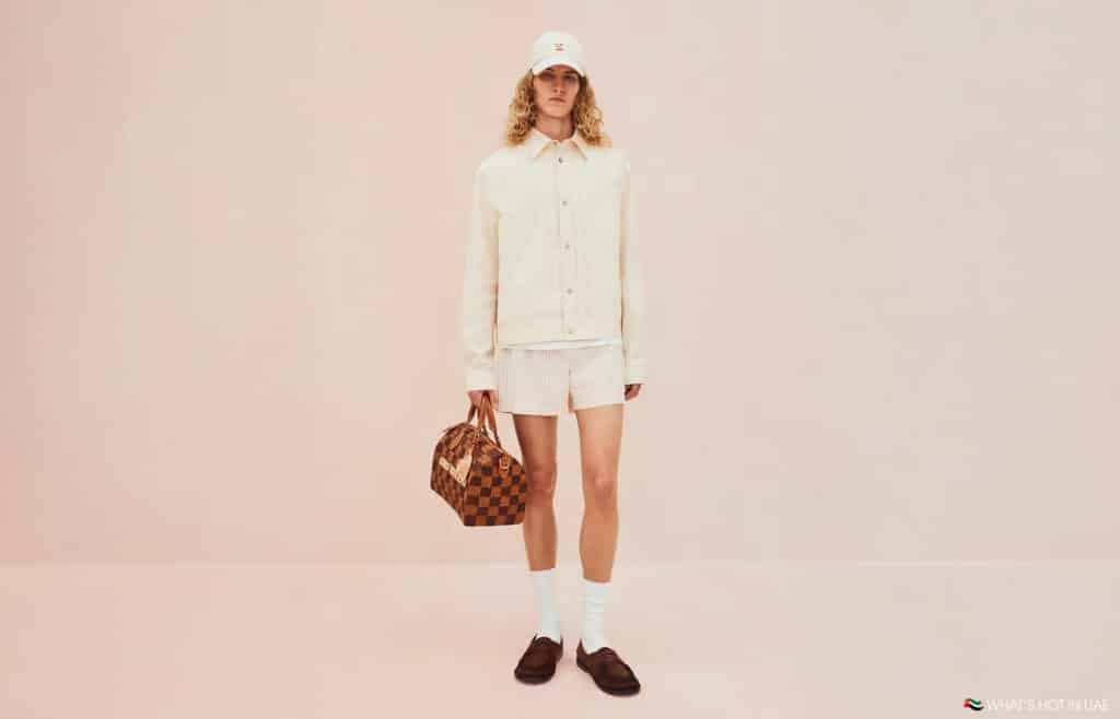 A person wearing a white long-sleeve shirt, white shorts, and a white cap, holding a brown Louis Vuitton Pre-Fall 2026 checkered bag, standing against a light pink background.