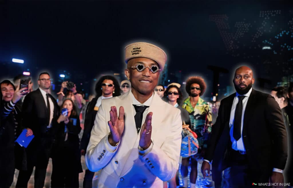 A person in a light-colored suit and beret claps while surrounded by a crowd, some of whom are taking photos, against a nighttime city backdrop with lights.