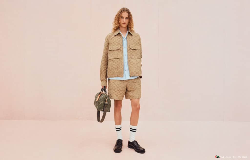 Person with long curly hair wearing a tan patterned jacket and shorts, holding a Louis Vuitton Pre-Fall 2026 branded bag, standing against a light pink background.