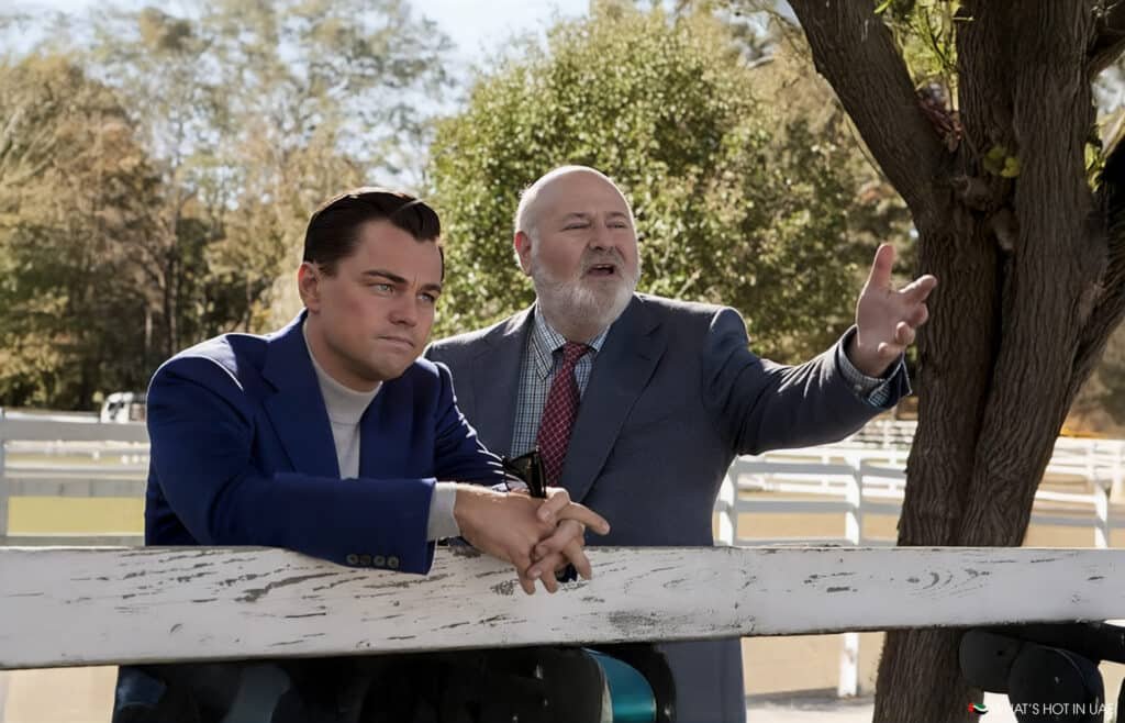 Two men in suits standing outdoors, one leaning on a white fence with a thoughtful expression, while the other gestures animatedly. The background features trees and a bright, sunny atmosphere.
