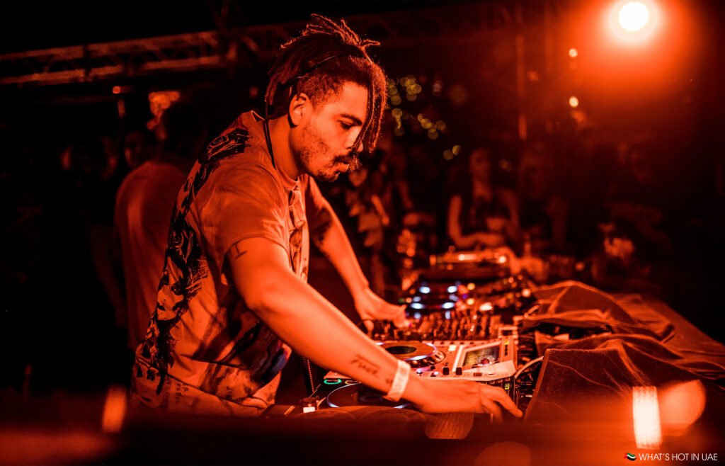 Seth Troxler is performing at a lively event under red lighting, engaging with his equipment on stage.