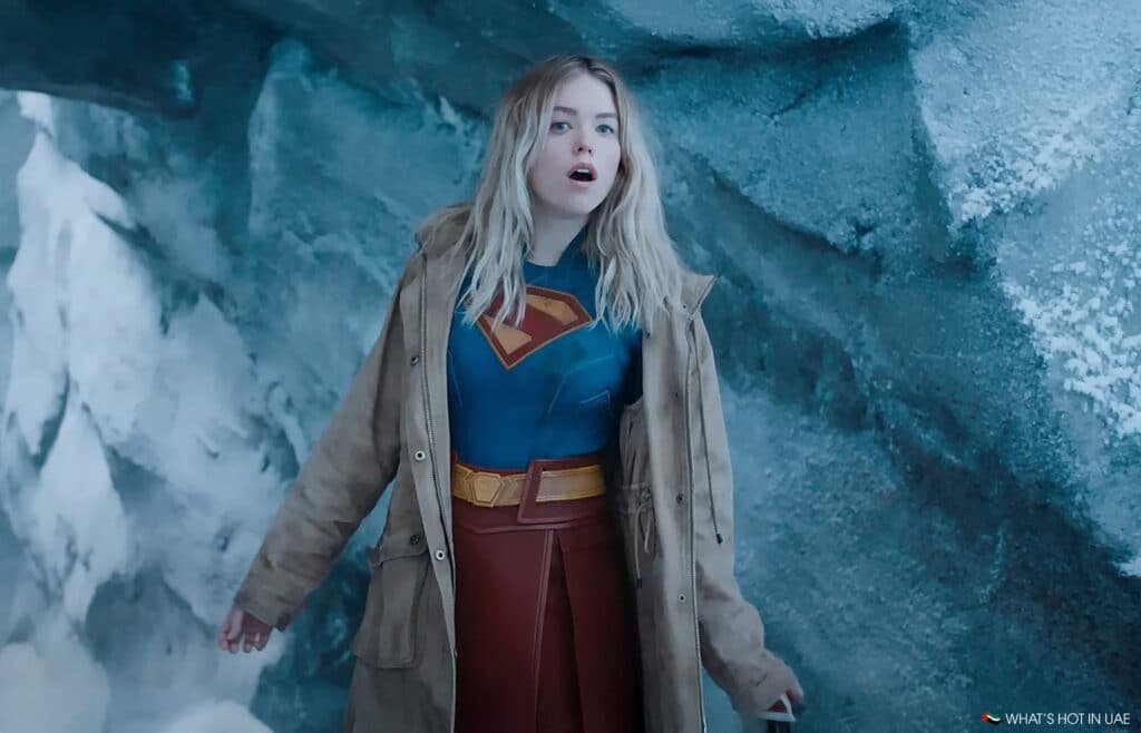 A woman wearing a Supergirl costume with a blue and red outfit featuring a large "S" emblem on her chest. She is standing in an icy, snowy environment, wearing a brown coat.