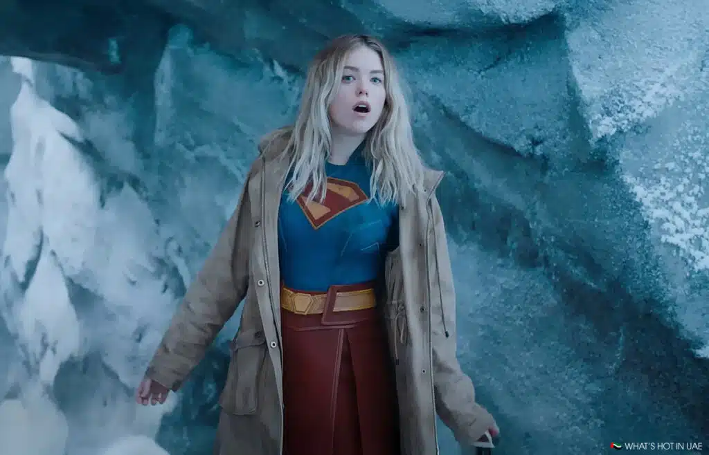 A woman wearing a Supergirl costume with a blue and red outfit featuring a large "S" emblem on her chest. She is standing in an icy, snowy environment, wearing a brown coat.