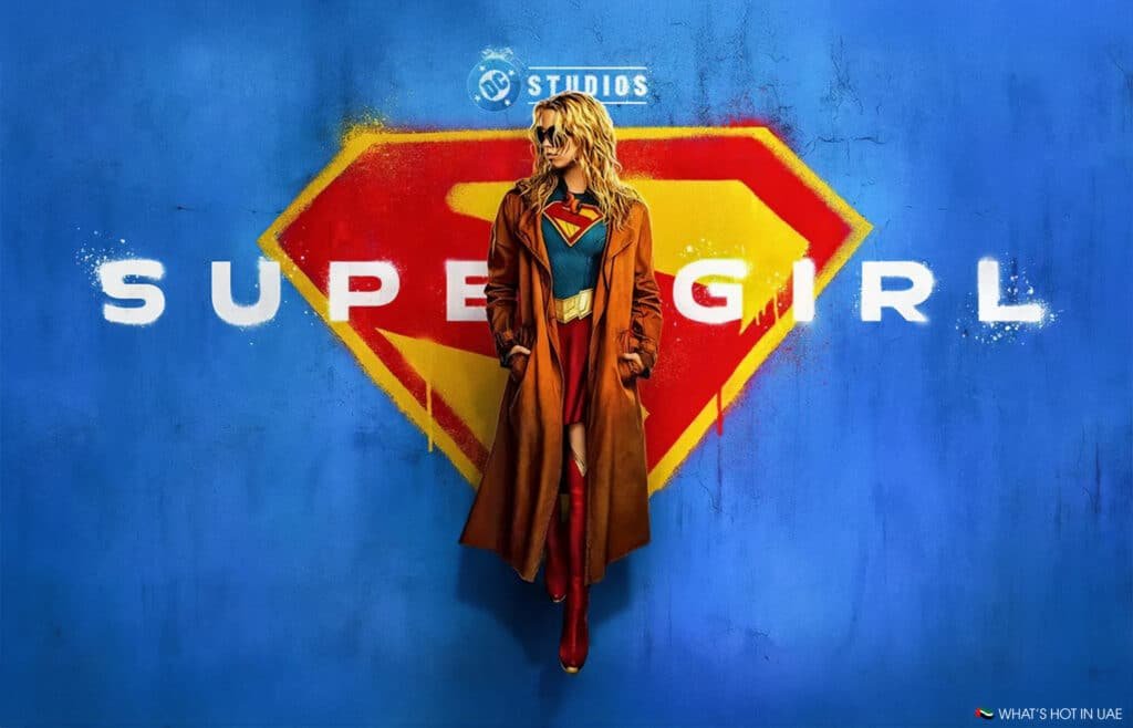 A character dressed as Supergirl, wearing sunglasses and a brown coat over a superhero costume, stands against a blue wall with a large Supergirl logo and the word 'Supergirl' in white letters.