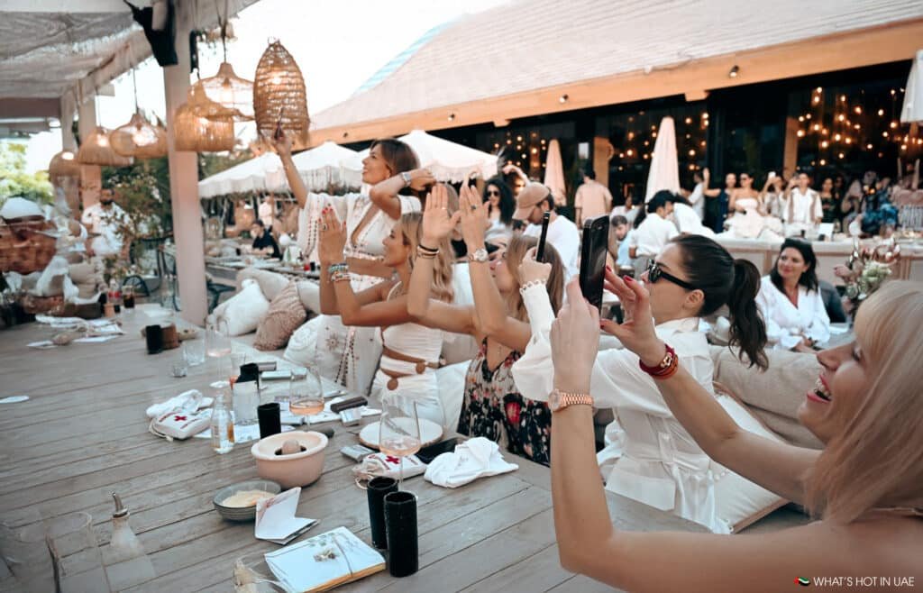 A group of people in a lively outdoor Hedonïsm restaurant setting, some taking photos with smartphones and others raising their hands, surrounded by cozy decor and hanging lights.
