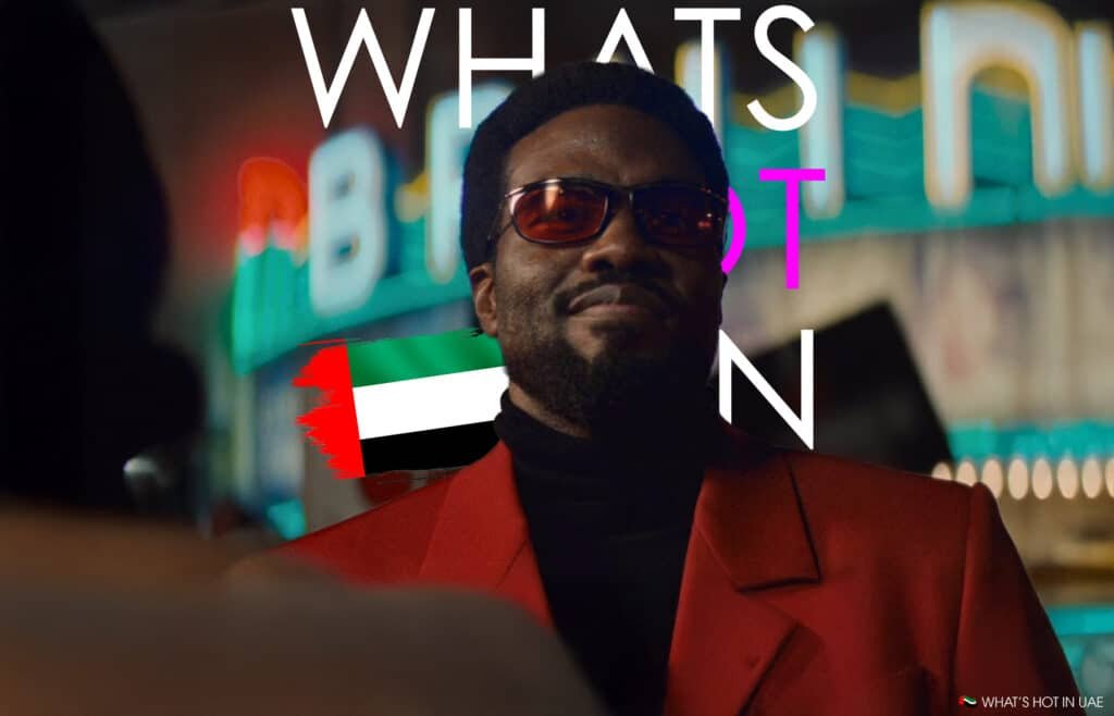 A person wearing sunglasses and a red suit stands in front of vibrant neon signs with "WHAT'S HOT IN UAE" written prominently.