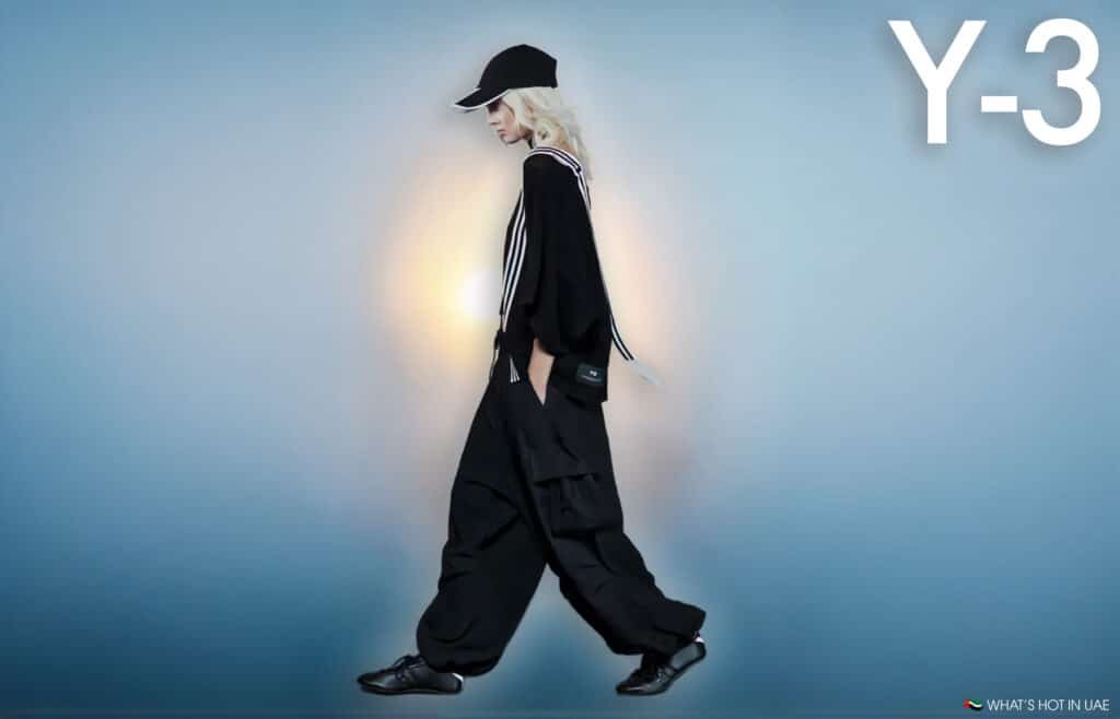 A person walking against a neutral background wearing a black outfit with oversized pants, striped shoulder details, and a black cap, featuring the Y-3 logo in the top right corner.
