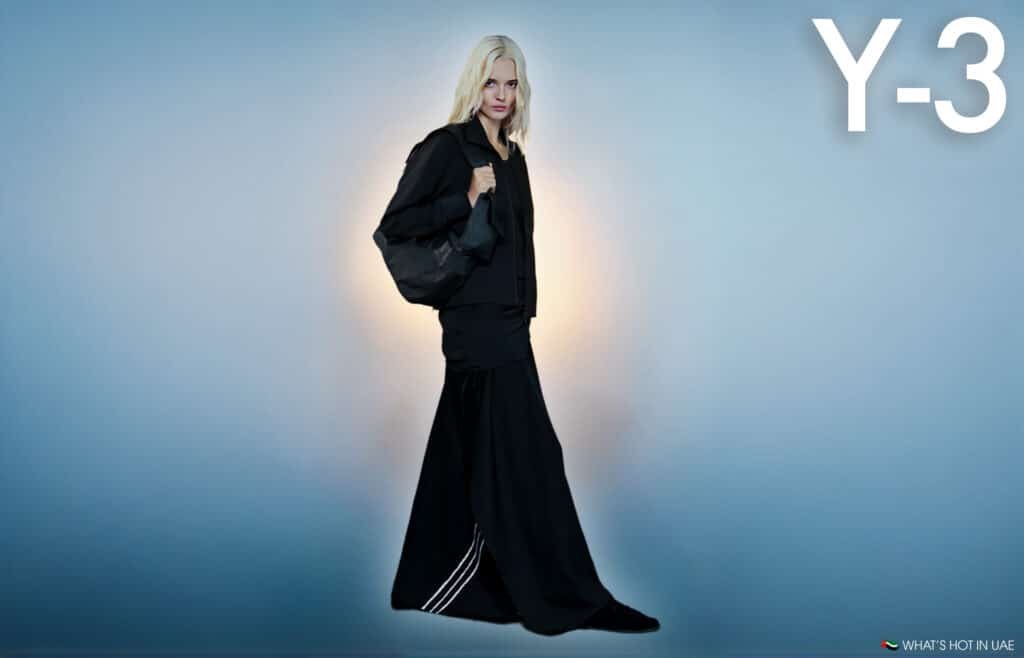 Person with blonde hair wearing a long black outfit and holding a black bag, with "Y-3" text displayed in the top right corner against a blue gradient background.