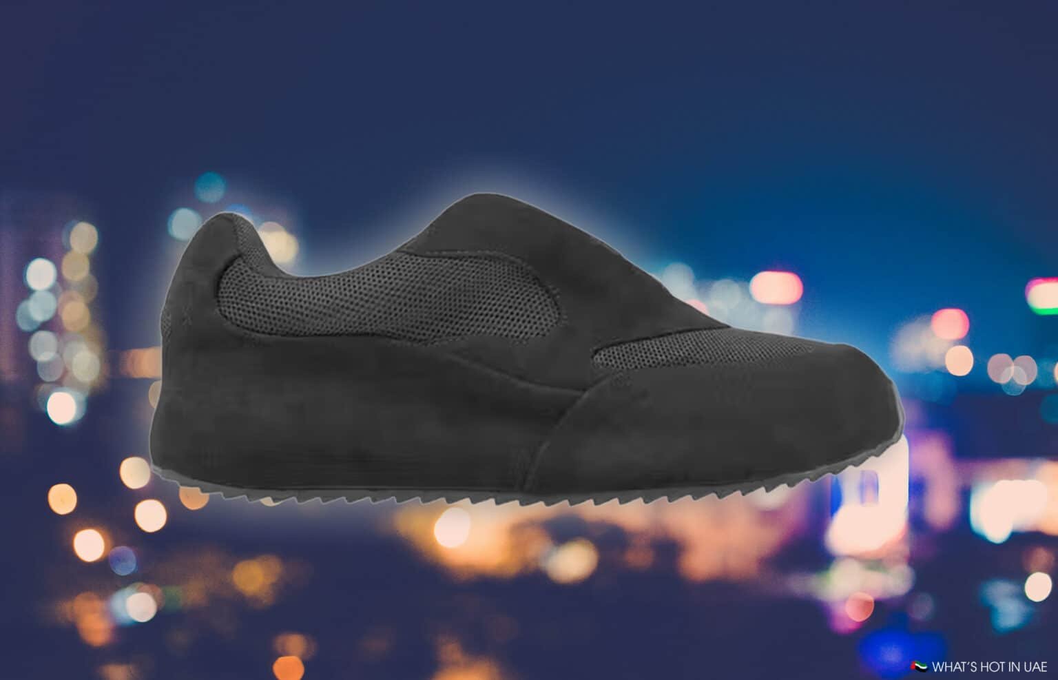 A black casual sneaker with a textured upper is set against a blurred nighttime cityscape.